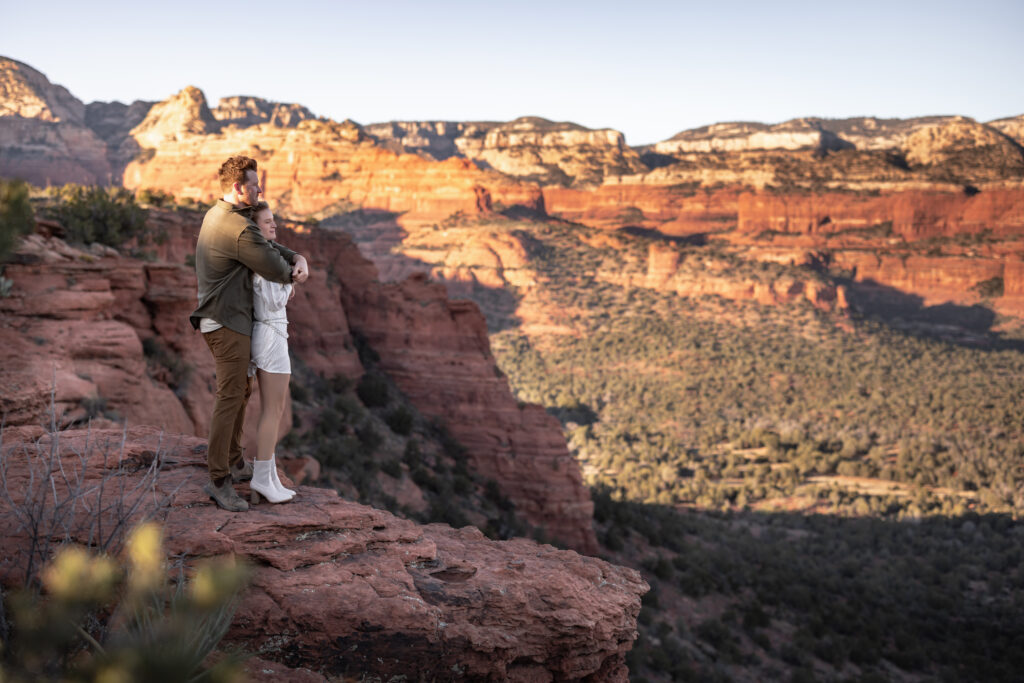 An engaged couple overlooking a cliff, holding each other in their arms over Sedona. A photo taken by Kollar Photography. Arizona Engagement photographer, Arizona Elopement Photographer. 