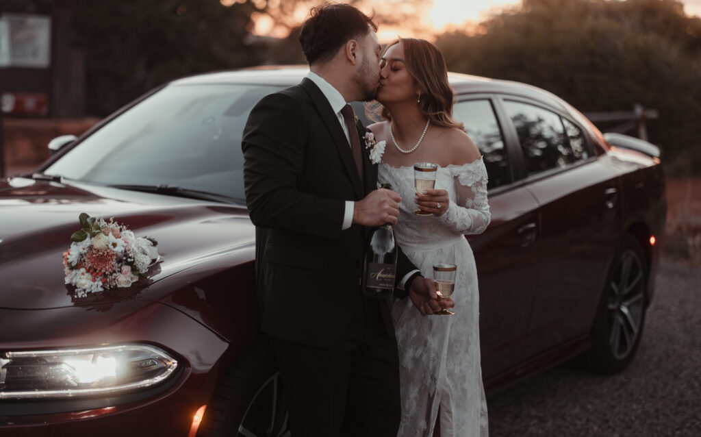 A bride and groom share a kiss while leaning against a burgundy car at sunset, holding champagne glasses, with a bouquet resting on the hood, creating an intimate destination wedding moment.