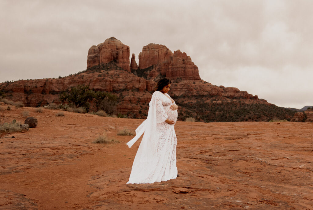 A pregnant woman wearing a flowing white lace dress stands gracefully on red rock terrain with the iconic Cathedral Rock of Sedona, Arizona, in the background. The overcast sky and dramatic landscape create a serene and majestic atmosphere.