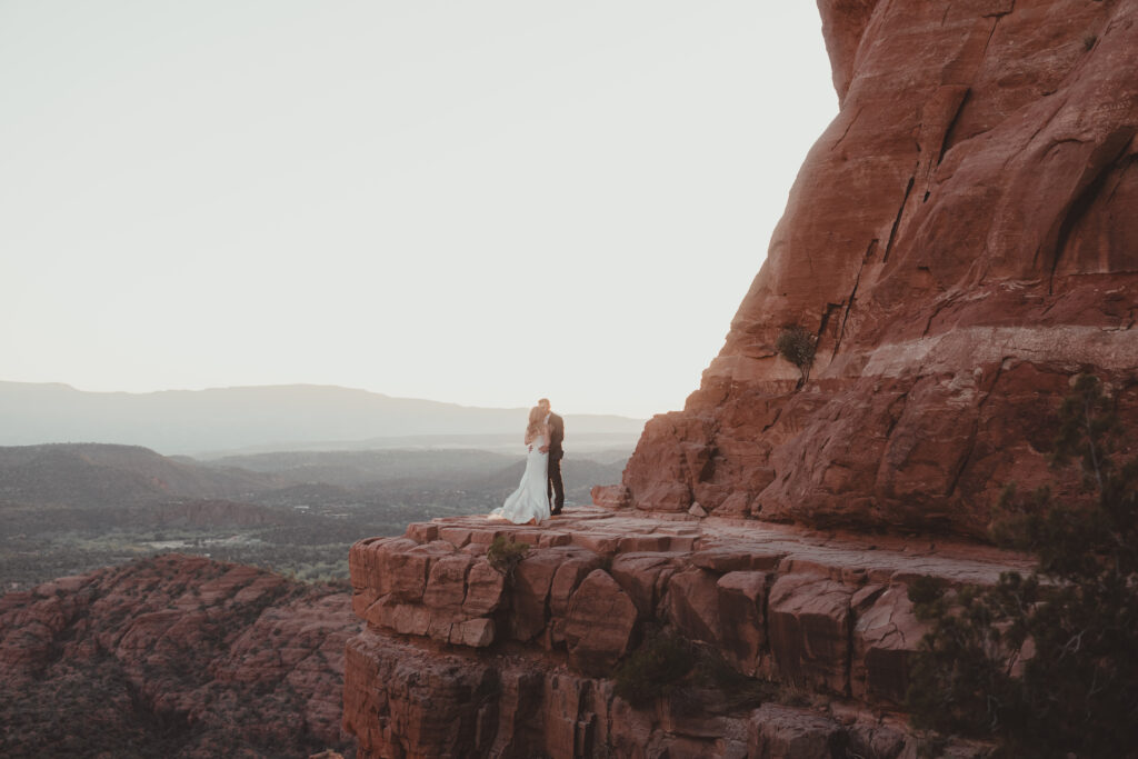 A bride and groom embracing on the edge of a red rock cliff at sunset, overlooking a breathtaking valley in Sedona, Arizona, during a destination wedding.