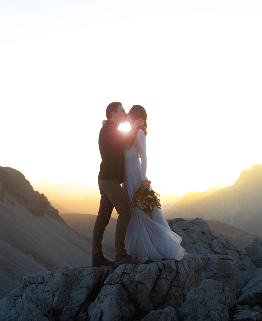 A bride and groom sharing a kiss on a rocky mountain peak at sunrise, with the bride holding a bouquet of autumn flowers and the golden light illuminating the dramatic landscape.