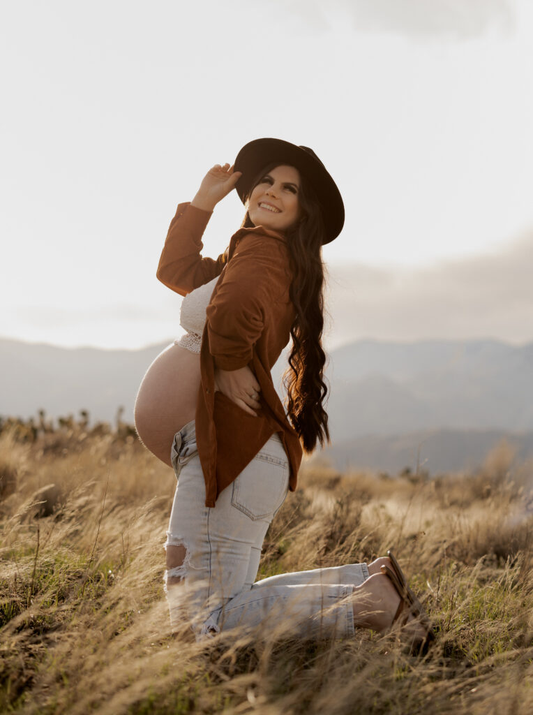 A glowing pregnant woman kneeling in a grassy field at sunset, wearing a lace top, an open brown jacket, and light-washed ripped jeans. She smiles brightly while holding her baby bump, with a black wide-brimmed hat adding a stylish, rustic touch. The soft golden light and mountains in the background create a serene and warm atmosphere.