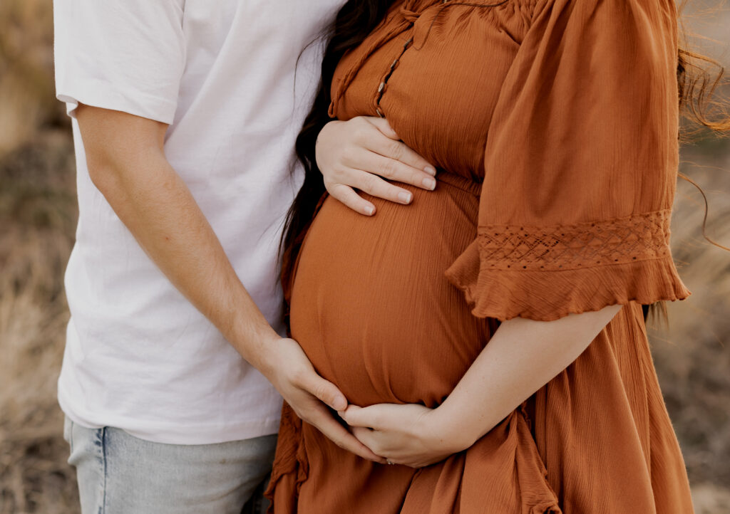 A close-up shot of a pregnant woman wearing a rust-colored flowy dress, with her partner’s hands gently placed over her baby bump, forming a heart shape. The natural outdoor setting adds warmth and intimacy to this tender moment.