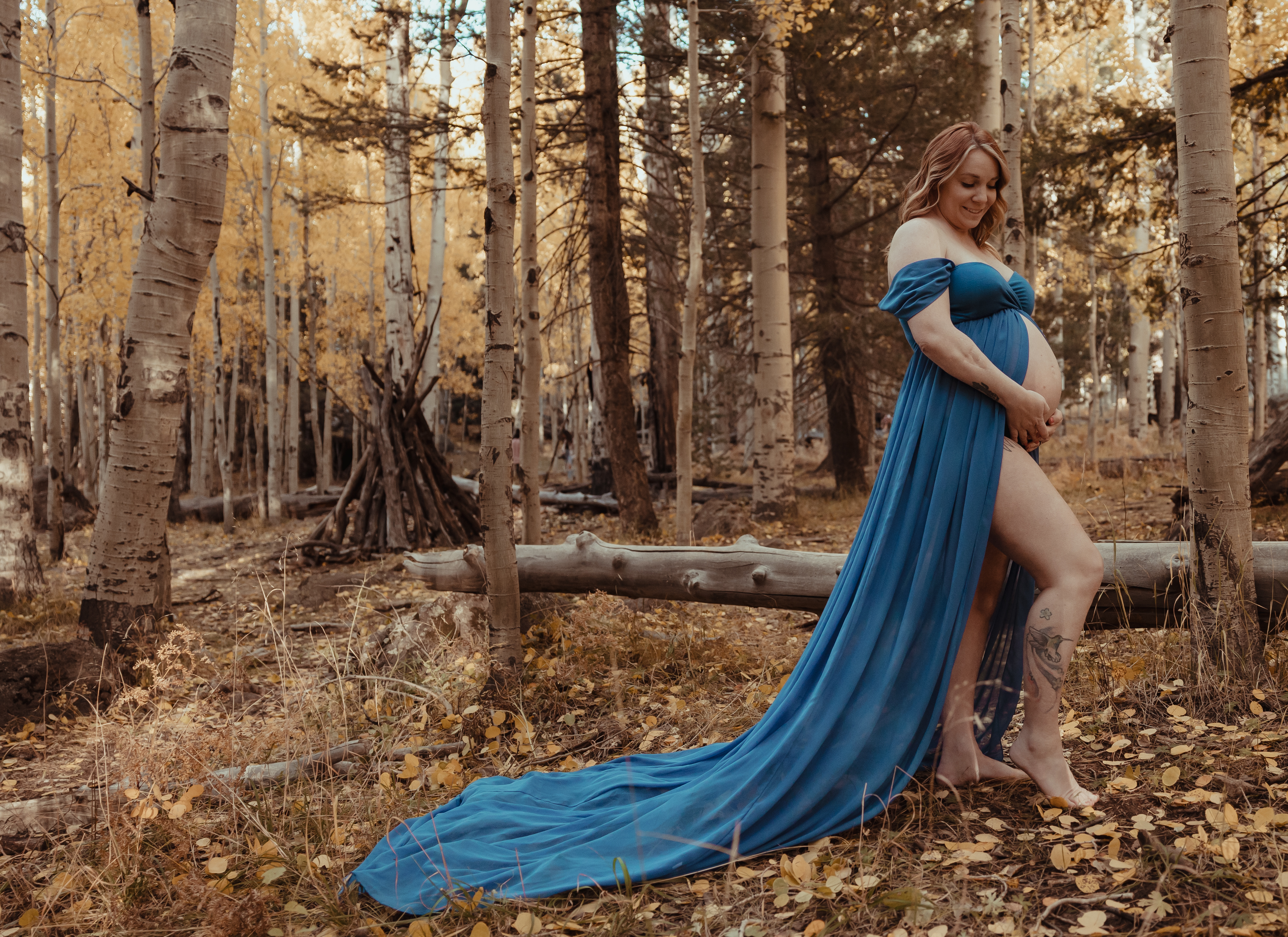 A pregnant woman wearing an off-shoulder flowing blue gown stands barefoot in a forest filled with golden autumn leaves and tall aspen trees. She gently cradles her baby bump while smiling, surrounded by a peaceful and rustic natural setting