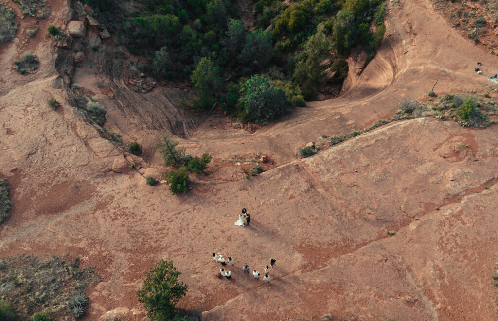 Aerial view of a small wedding ceremony taking place on a red rock formation, with the bride and groom standing together surrounded by guests, in a stunning desert landscape.