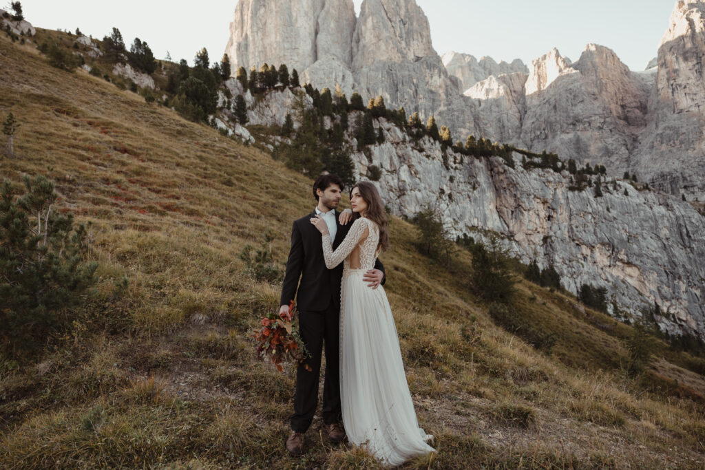 A bride and groom celebrating their destination wedding with a champagne toast at sunset, leaning against a stylish burgundy car adorned with a floral bouquet.